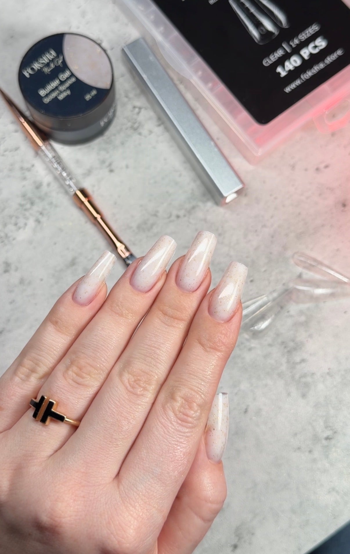 Hand with long, white nail polish small brush, with a container of nail gel and a box of false nails in the background.