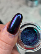 Nail polish with a metallic blue and purple finish on a finger, next to an open nail polish jar.