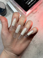 Hand with long, clear-coated nails holding a small container on a light background