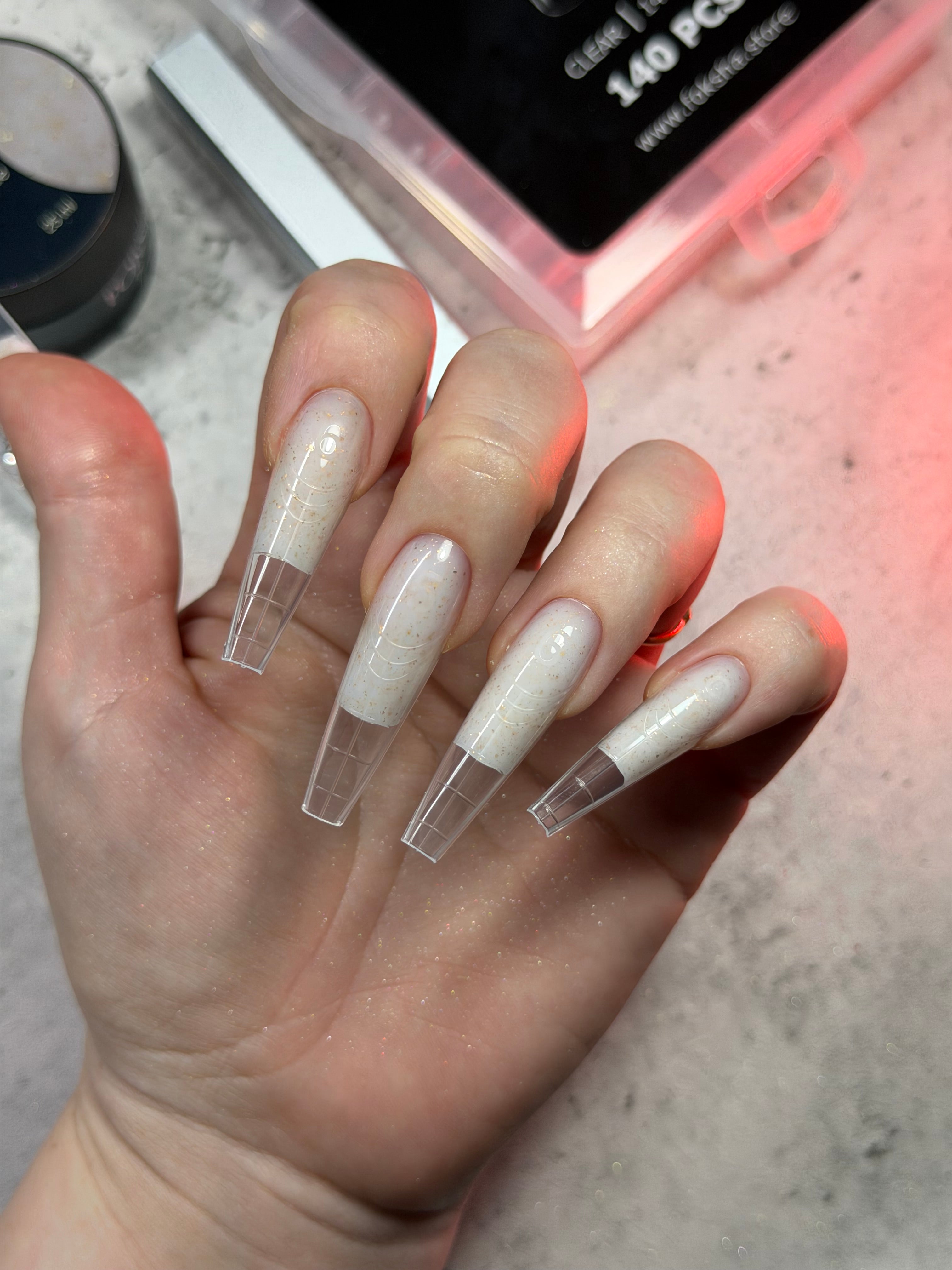 Hand with long, clear-coated nails holding a small container on a light background