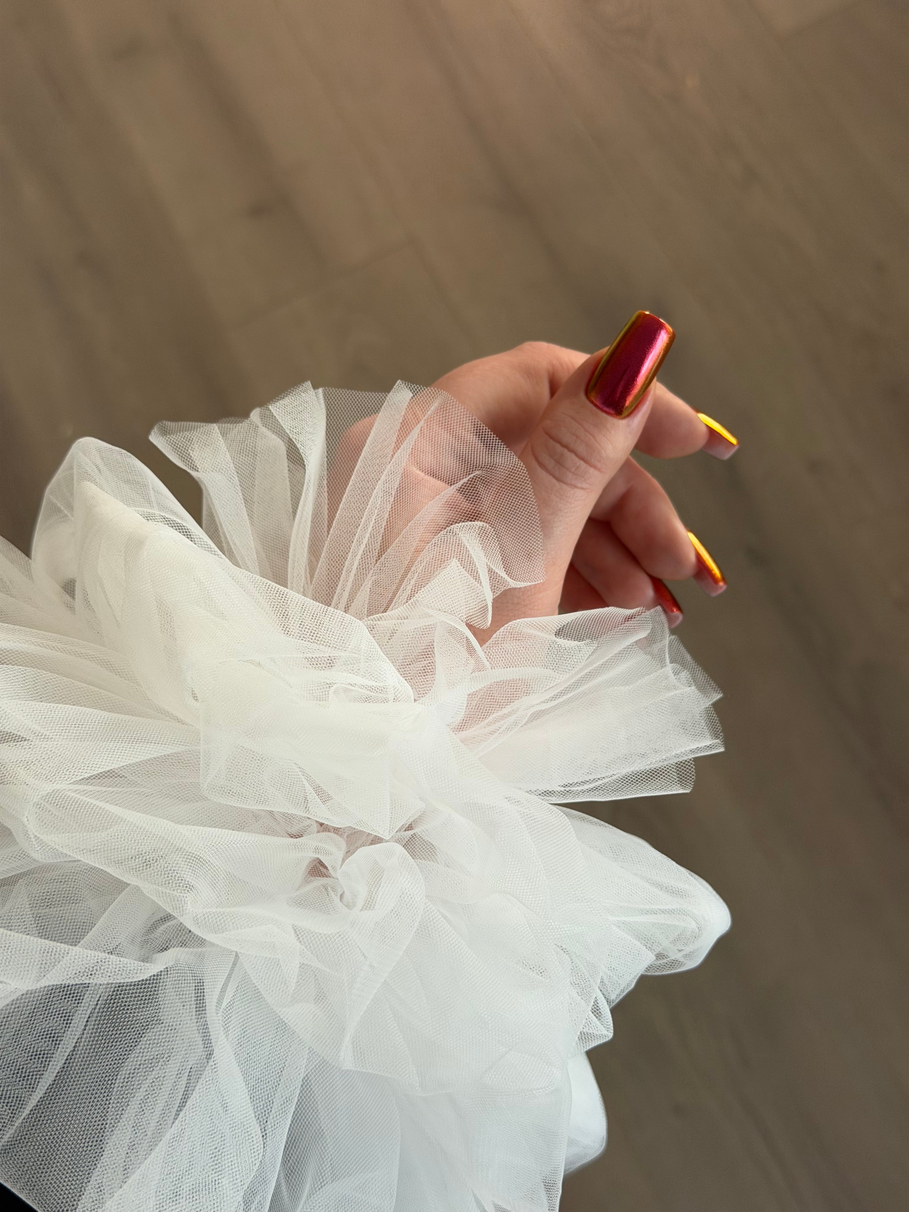 Close-up of a hand with red nail polish holding a white tulle fabric against a wooden floor background.